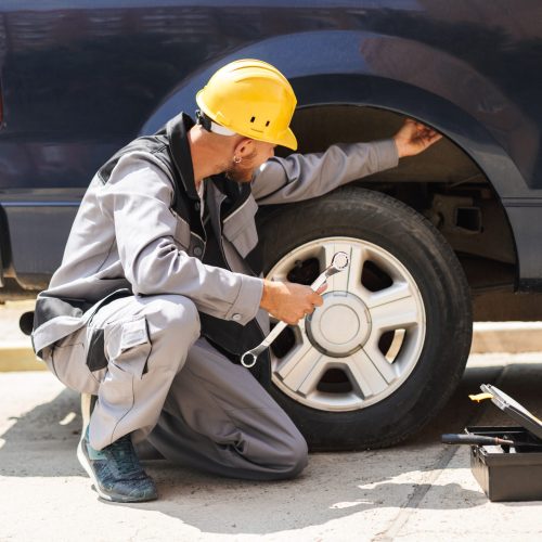 Young mechanic in work clothes and yellow hardhat using wrench for changing car wheel outdoor